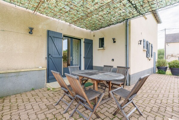 En patio med et rundt bord og stole, et overdækket område med en espalier-tag, og en bygning med beige facade og blå skodder.