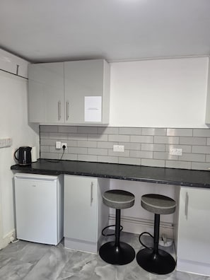 A kitchen with white cabinets, a black countertop, a small white refrigerator, and two black stools. - City Centre, P/ Bathroom,room-7, 
City View Place (Cambridge)