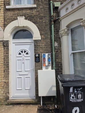 A white door with a small window above it, a utility box with numbered buttons, and a black trash bin with a logo. - City Centre, P/ Bathroom,room-7, 
City View Place (Cambridge)