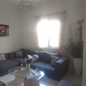 A living room with a sofa, a glass coffee table, a vase with flowers, and a large window with curtains.