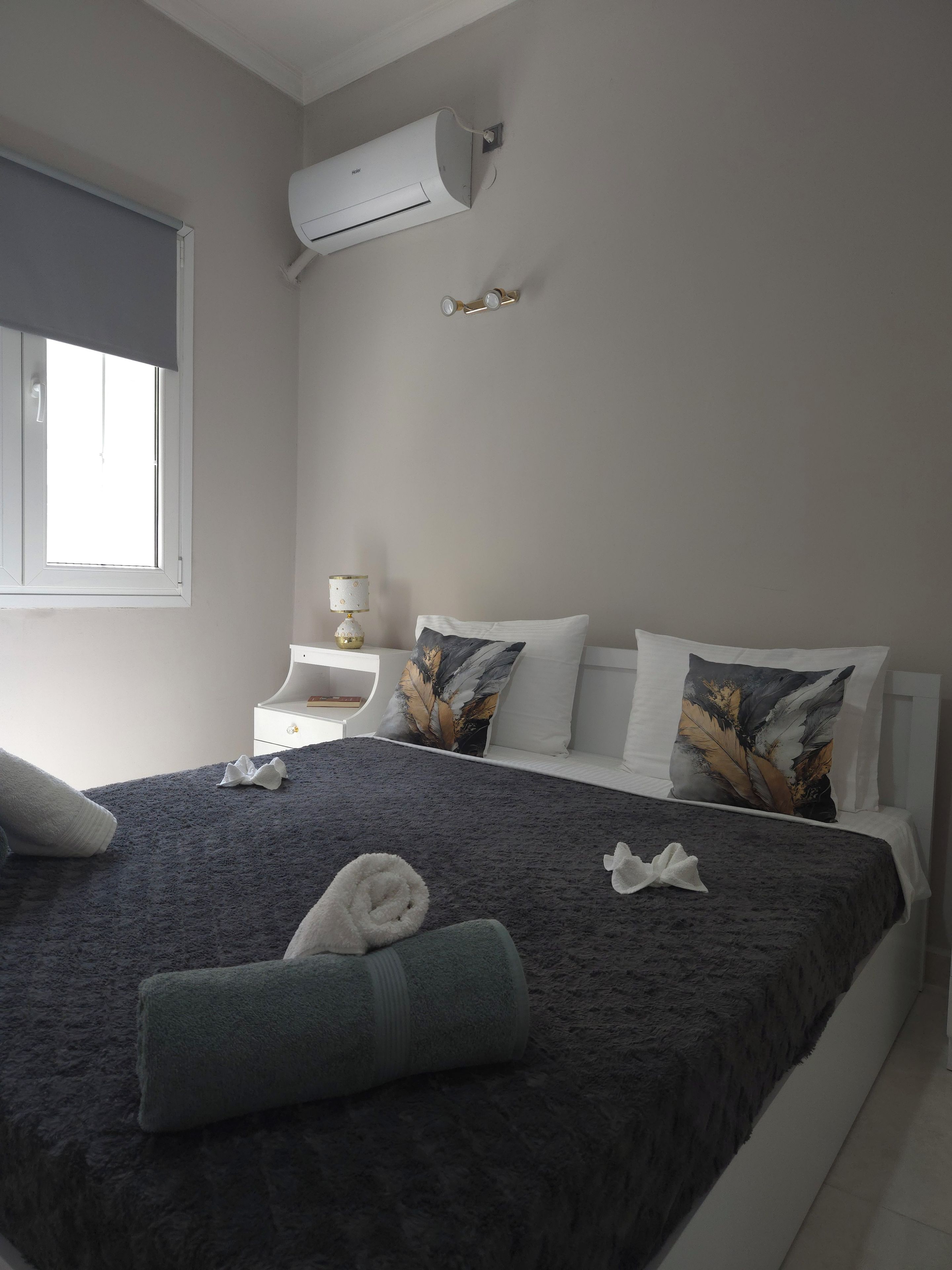 Una habitación de hotel con cama, almohadas, mesita de noche, lámpara y un equipo de aire acondicionado.