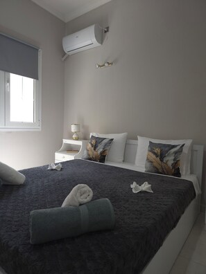 A hotel room with a bed, pillows, a nightstand, a lamp, and an air conditioning unit. - Enora Loutraki! Stylish 2BR Retreat with private Garden near the Sea. (Loutraki)