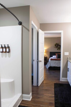 Baño con ducha, inodoro y lavamanos. Hay un pasillo que conduce a un dormitorio con cama y mesita de noche.