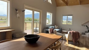 A spacious living area with a wooden table, a sofa, and a view of a mountainous landscape through large windows.