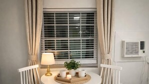 A corner room with a window, white curtains, a table with chairs, a lamp, a potted plant, and an air conditioning unit. - Salt Air Suites • king bed • Pool• Pets OK #2 (Clearwater)