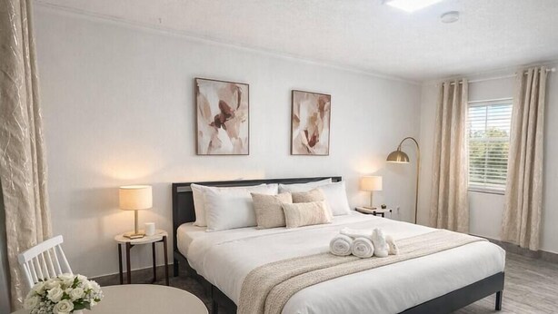 A hotel room with a bed, two wall-mounted lamps, a round table with chairs, and a window with curtains. - Salt Air Suites • king bed • Pool• Pets OK #2 (Clearwater)