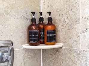 Drei Flaschen Shampoo und Conditioner stehen auf einem weißen Regal in einem Badezimmer mit einem Wandbelag aus gesprenkelten Fliesen.