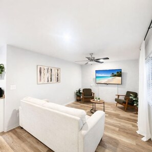 Ein Wohnzimmer mit einer Couch, zwei Sesseln, einem Couchtisch, einem Deckenventilator, einem Fernseher und einem Bild an der Wand.