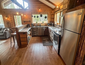 A cabin-style kitchen with wooden walls, a central island, stainless steel appliances, and a large window overlooking greenery.