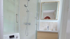 Casa de banho compacta com cabine de duche em vidro, máquina de lavar roupa, lavatório com espelho e um pequeno contentor de lixo.
