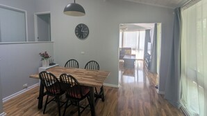 A dining area with a wooden table, four chairs, a black pendant light, a wall clock, and a view through a doorway to another room.