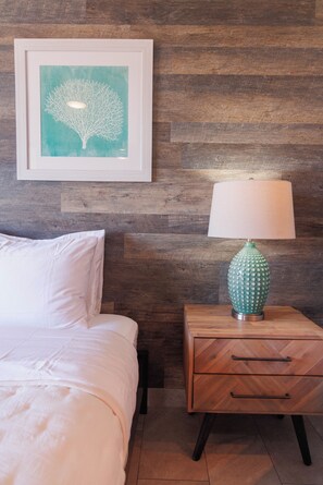 A neatly made bed with white bedding, a wooden nightstand with a green lamp, and a framed artwork on the wall. - Holo Hou Hale - Lahaina Shores Retreat on Front St (Lahaina)