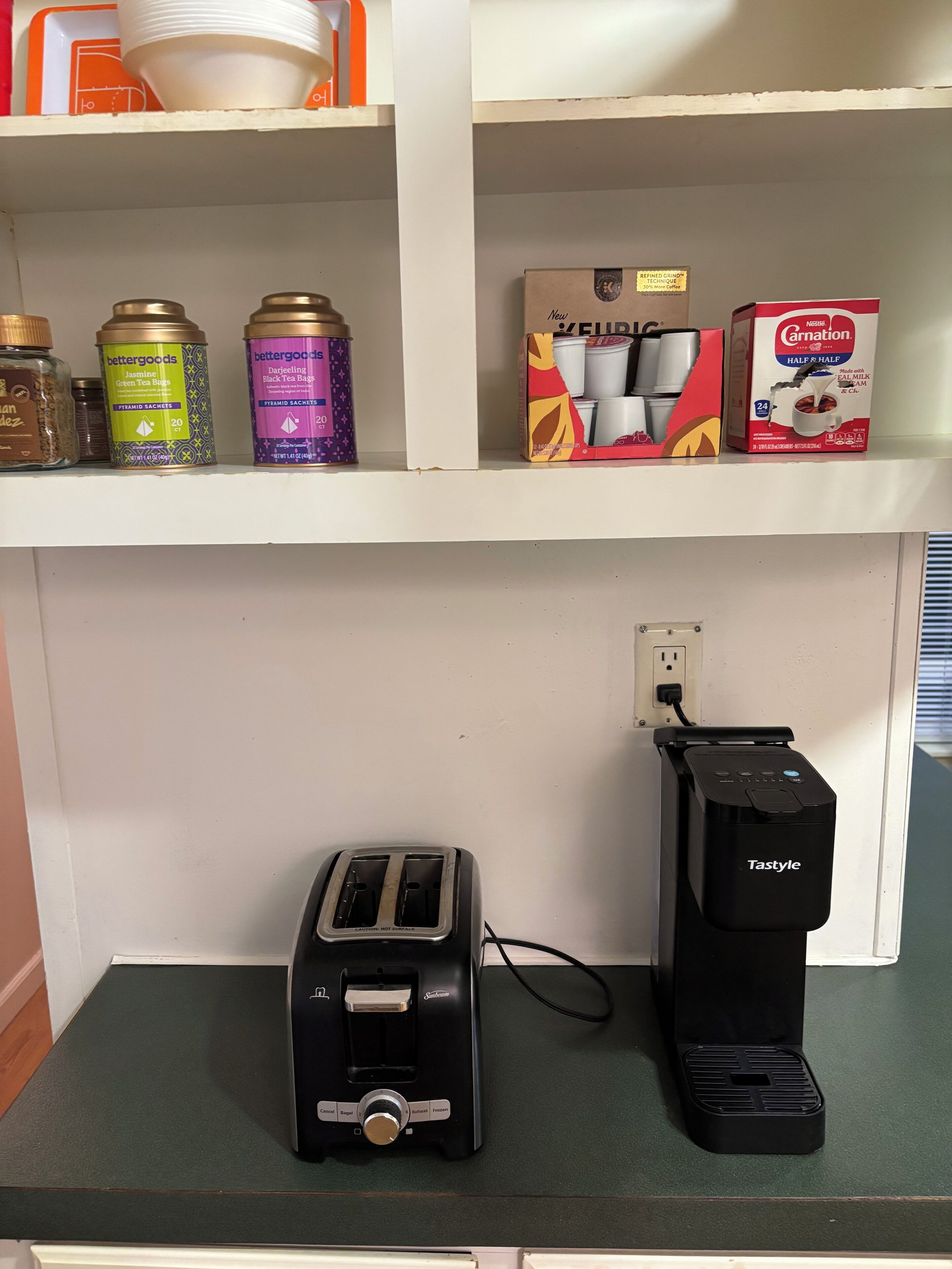 Una cocina con un tostador y una cafetera en la encimera, y repisas superiores con bolsitas de té y una caja de cereales.