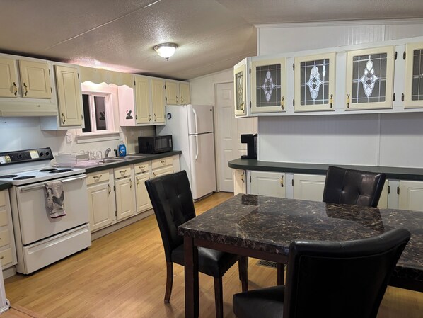 Una cocina con armarios blancos, encimera negra, estufa, refrigerador y una mesa de comedor con sillas.
