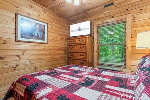 Quarto de cabana de madeira, com uma cama, cômoda, quadro na parede e janela com persianas.