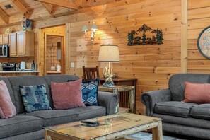 Interior acolhedor de uma cabana, com teto de madeira, mesa de jantar, conjunto de sofás e área de cozinha.