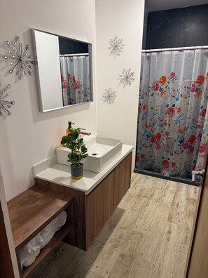 Ein Badezimmer mit weißem Waschbecken, Holz-Waschtisch, einer Topfpflanze, Wanddekoration und einer Duschvorhang mit floralem Muster.