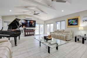 A spacious living room with a grand piano, a glass coffee table, a comfortable sofa, and a ceiling fan. - Captain's Cove | Canalfront w/ Pool & Boat Lift (Key West)