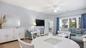 Ein modernes Wohnzimmer mit einem runden weißen Tisch, weißen Stühlen, einem grauen Sofa, einem Flachbildfernseher und einem Deckenventilator.