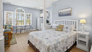 Ein Schlafzimmer mit Bett, Schreibtisch, Stuhl, einem Fenster mit Jalousien, einem Kühlschrank und einer Lampe.