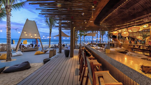 En bar vid stranden med sittplatser på ett trädäck, en bardisk och utsikt över havet.