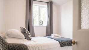 A bedroom with a bed, pillows, a window with curtains, a clock, and a door. - 2 Crossing Cottage, Theberton (Leiston)