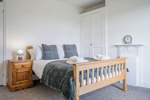 A bedroom with a wooden bed, a nightstand with a lamp, a dresser, a white wardrobe, and a fireplace mantel with a clock. - 2 Crossing Cottage, Theberton (Leiston)