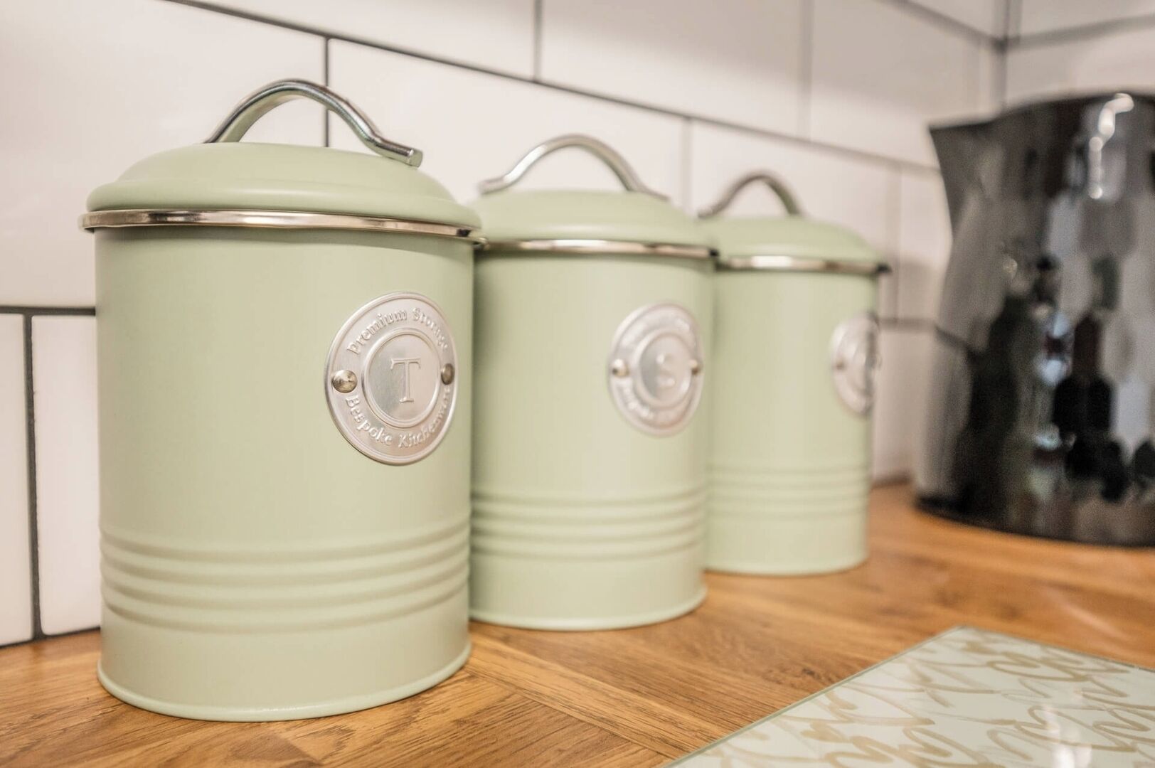Three green kitchen storage containers with metal handles and lids, placed on a wooden countertop against a tiled backsplash.