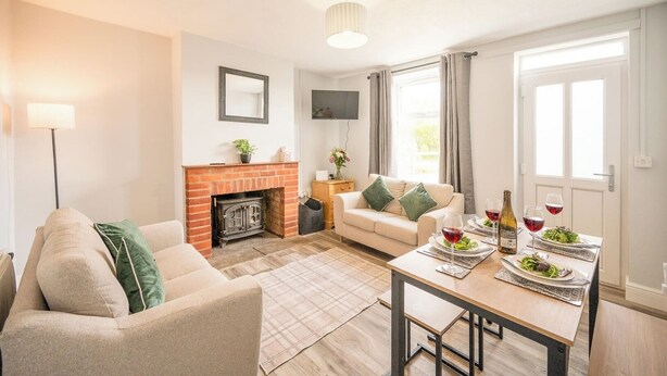 A living room with a fireplace, sofa, table with dinner set, and a TV mounted on the wall. - 2 Crossing Cottage, Theberton (Leiston)