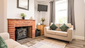 A living room with a brick fireplace, a beige sofa with green cushions, a television mounted on the wall, and a window with curtains. - 2 Crossing Cottage, Theberton (Leiston)