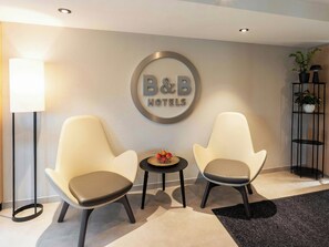 A hotel lobby with two chairs, a small table with fruit, a lamp, and a sign for B&B Hotels.