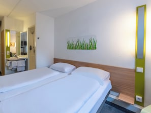 Quarto de hotel com uma cama, banheiro com espelho e luminária fixada na parede.
