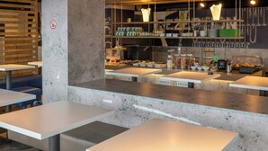 A modern cafe interior with white tables, a concrete pillar, and a counter with various food items and utensils. - B B Hotel Braunschweig West (Braunschweig)