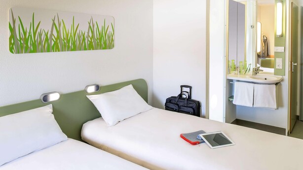 A hotel room with two beds, a bedside table with a tablet, a suitcase, and a bathroom with a sink and mirror. - B B Hotel Braunschweig West (Braunschweig)