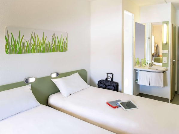 Quarto de hotel com duas camas, uma mesinha de cabeceira com um tablet, uma mala e um banheiro com pia e espelho.