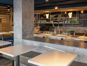 Interior de una cafetería moderna con mesas blancas, un pilar de hormigón y una barra con diferentes alimentos y utensilios.