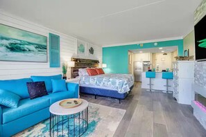 Une chambre moderne avec un canapé bleu, un lit avec une literie à motifs, une petite table ronde et un coin cuisine avec un bar.
