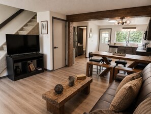 A modern living room with a TV, wooden furniture, a staircase, and a kitchen in the background.
