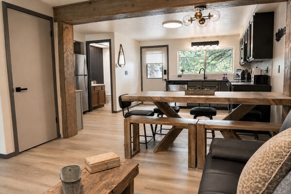 A modern kitchen with a wooden table and chairs, a dining area with a bench, and a view of the outdoors through large windows.