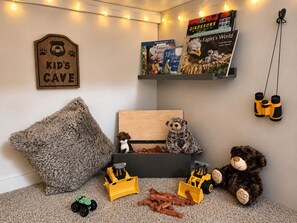 Un área de juegos para niños con un letrero que dice 'Kids Cave', libros, peluches y vehículos de juguete.