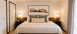 A bedroom with a large bed, two bedside tables with lamps, a framed picture on the wall, and a door on the left.