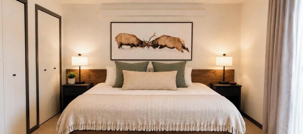 Un dormitorio con una cama grande, dos mesitas de noche con lámparas, un cuadro en la pared y una puerta a la izquierda.