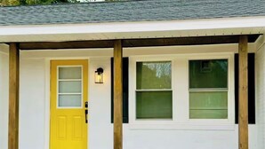 A house with a yellow front door, a porch with wooden posts, and two windows with blinds. - Elton Home (Goldsboro)