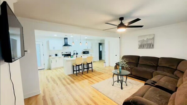 A modern living room with a sofa, a TV, a ceiling fan, and a kitchen in the background. - Elton Home (Goldsboro)