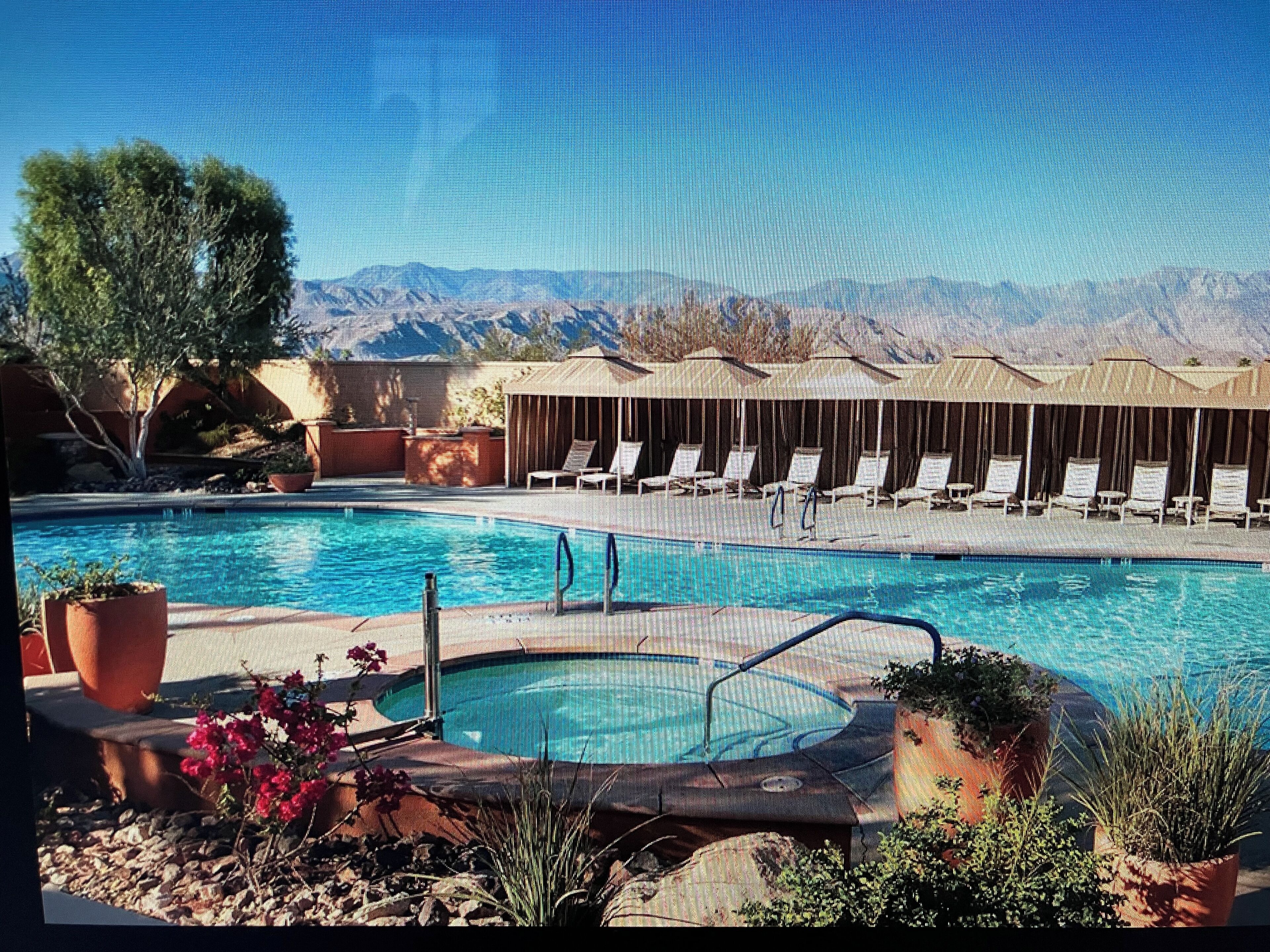 A pool with lounge chairs and a hot tub, surrounded by potted plants and a mountain backdrop.