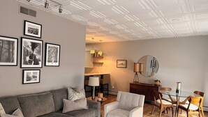 Una sala de estar moderna con un sofá gris, un sillón y una mesa de comedor con sillas. Hay cuadros enmarcados en la pared, una lámpara y un espejo.