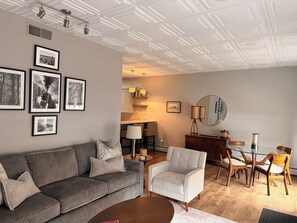A modern living room with a grey sofa, armchair, and dining table with chairs. There are framed pictures on the wall, a lamp, and a mirror.
