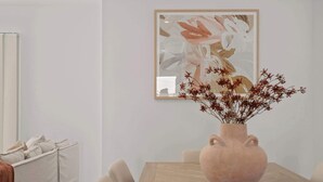 A dining area with a wooden table, a vase with dried flowers, and a framed abstract painting on the wall.