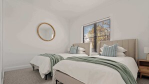 A bedroom with two beds, a round mirror, a window with blinds, and a bedside table with a lamp.
