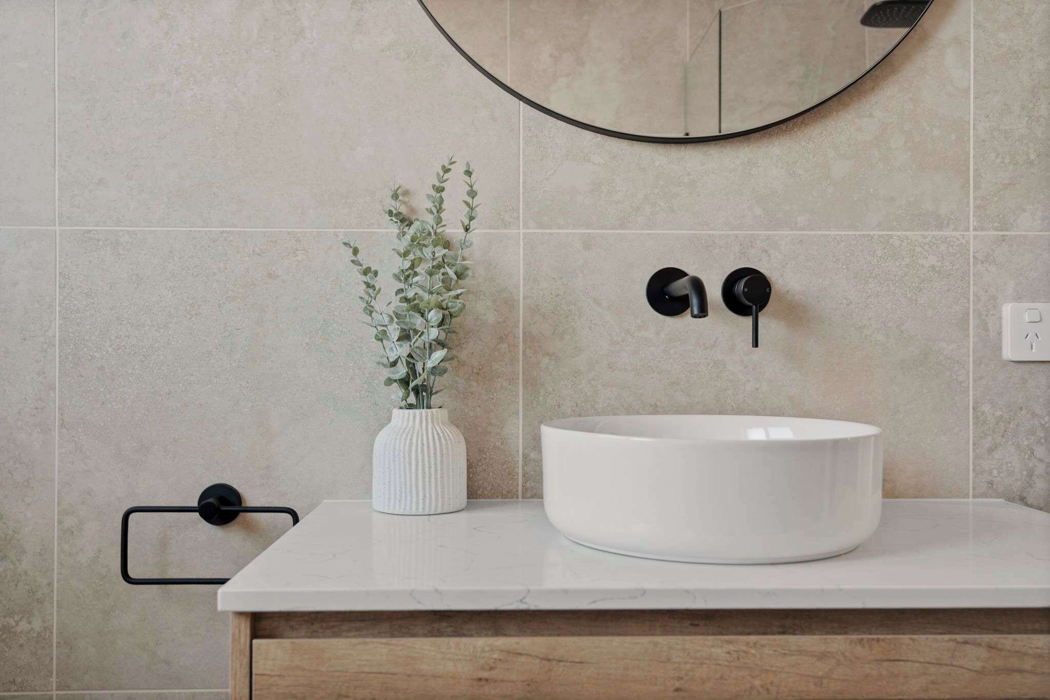 Un baño con un lavamanos blanco, un espejo redondo, un pequeño florero blanco con plantas verdes y un toallero negro.